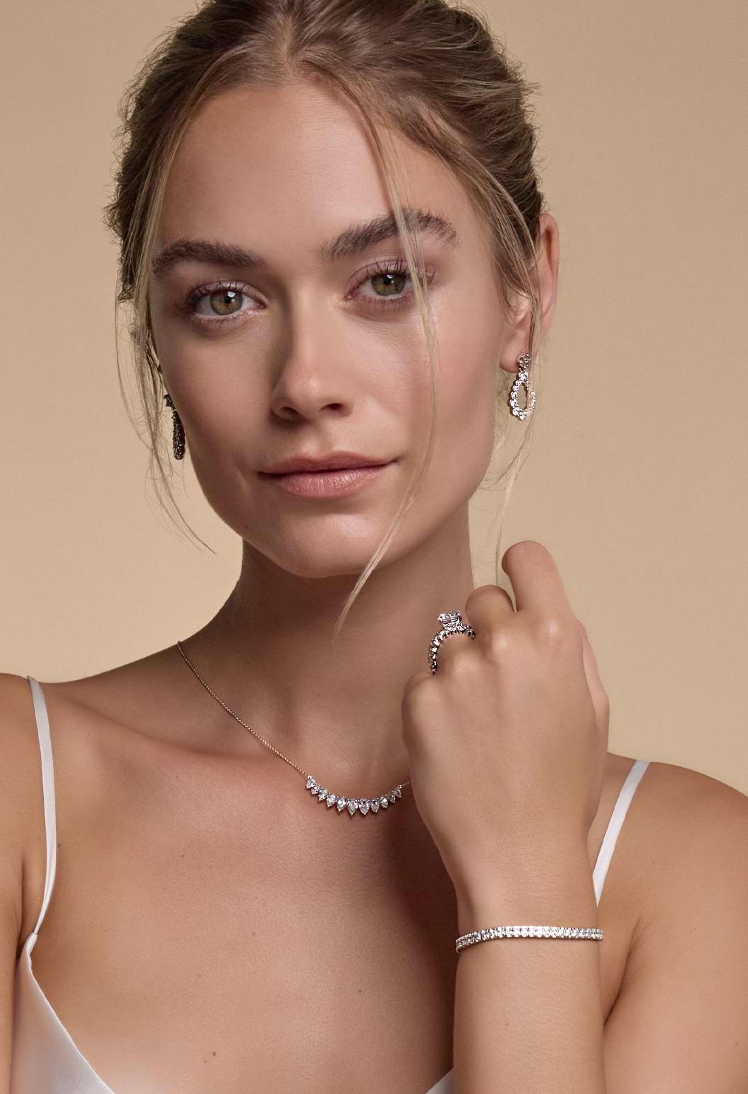 A closeup image of a female model looking at the camera showcasing diamond drop earrrings, necklace, tennis bracelet, enternity band and engagement ring