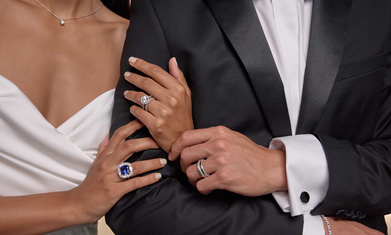 Image of intertwined hands of a bride and groom featuring a diamond pendant, fashion ring, eternity band and engagement ring on the bride's hands.The groom's hand features one men's wedding band. 