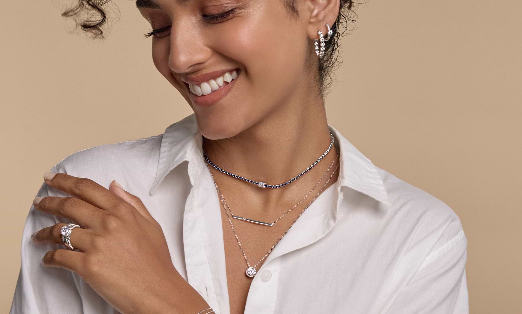 A side profile of a female model smiling with two diamond hoops, three fashion necklaces, an enternity band and an engagement ring