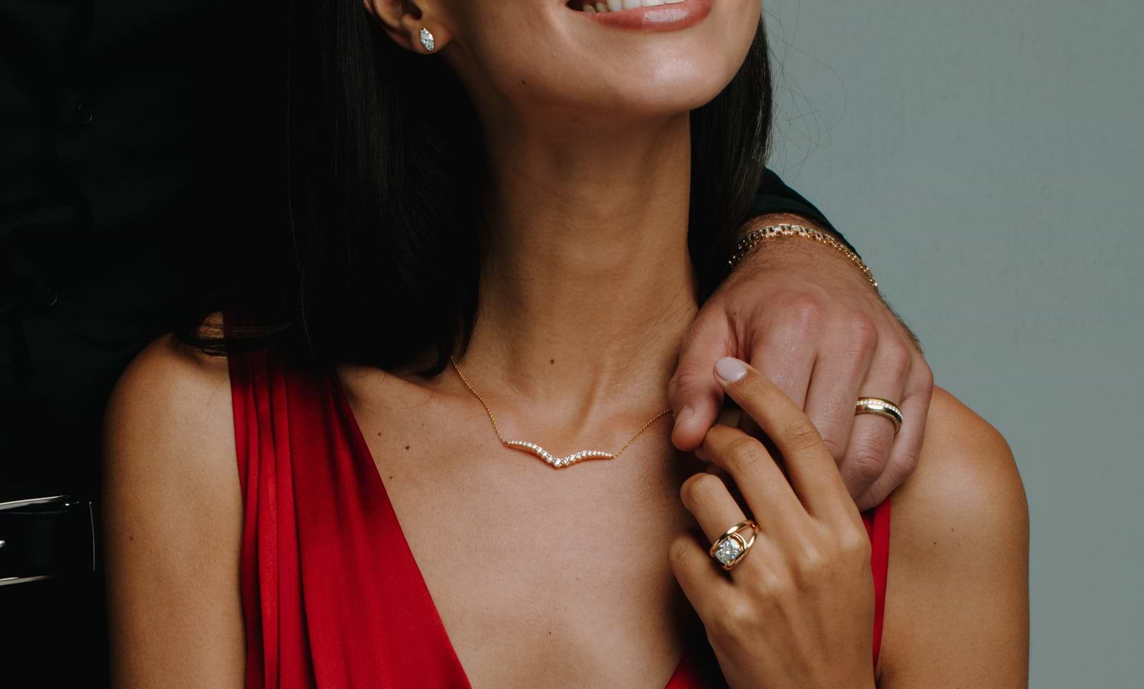Image of intertwined hands of a bride and groom featuring a diamond pendant, fashion ring, eternity band and engagement ring on the bride's hands.The groom's hand features one men's wedding band. 